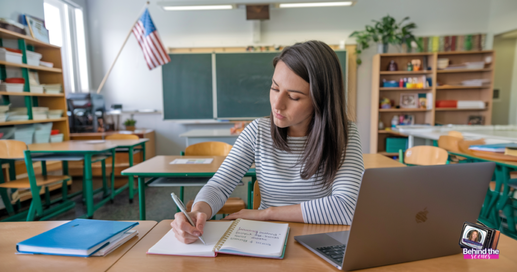 A woman with long hair, wearing a striped shirt, sits at a desk in a classroom. She is writing in a notebook with a laptop open beside her. The room has wooden desks and chairs, a blackboard, shelves, and a U.S. flag in the background.
