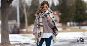 A person in a cozy sweater, jeans, and a large plaid scarf walks outdoors on a chilly day. Snow patches dot the ground, with bare trees and distant buildings in the background.