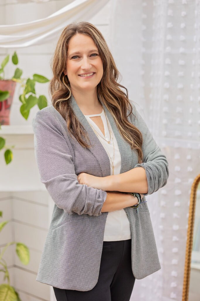 A woman with long brown hair stands indoors, smiling with arms folded. She wears a light gray blazer over a white top and black pants. Green plants and sheer white curtains are in the background.