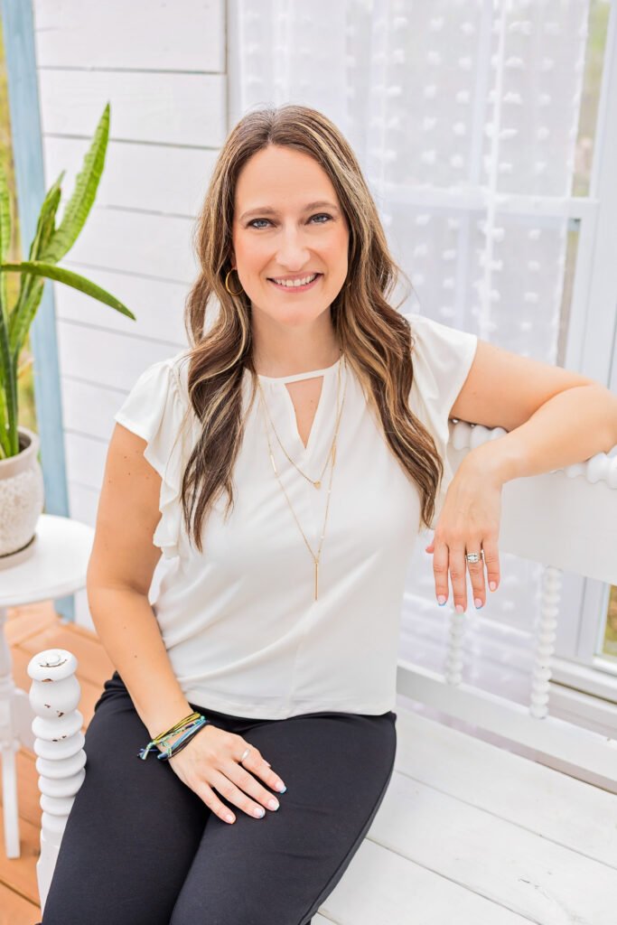 A woman with long brown hair and a white blouse sits on a white bench, smiling at the camera. She wears black pants, layered necklaces, and bracelets. A potted plant is in the background near a window with sheer curtains.