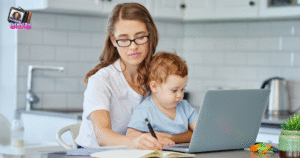 A woman wearing glasses follows a work from home schedule with kids, working on a laptop and writing in a notebook at a kitchen table while holding her toddler, who looks at the laptop screen in their bright, modern kitchen.