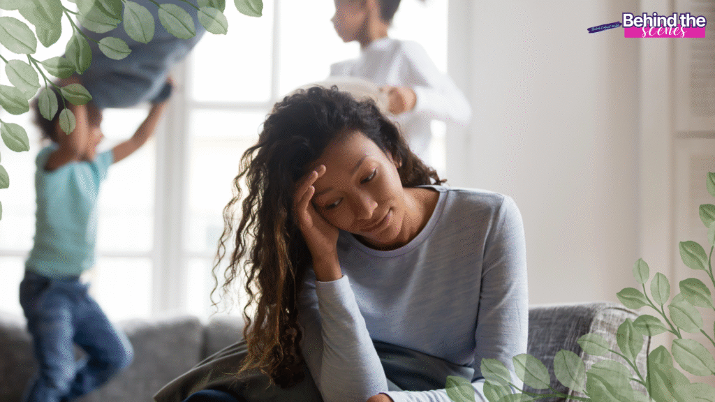 A tired woman sits on a couch with her hand on her forehead, looking down, capturing the challenge of reinventing yourself after kids. Two children play energetically in the background. Leaf graphics frame the image with "Behind the scenes" text in the top right corner.