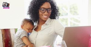 A woman with curly hair and glasses smiles while working on a laptop, holding a sleeping baby in her arms. Sunlight streams through a window behind them, and a logo in the top left reads Behind the View.
