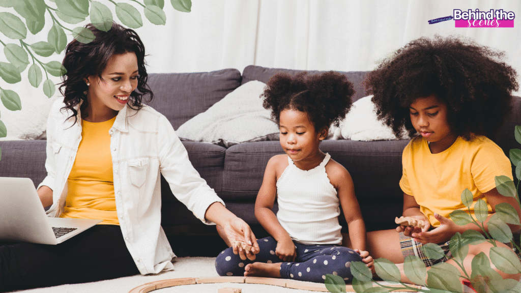 A woman sits on the floor with a laptop, smiling at two young girls who are playing with wooden toys. They are in a cozy living room with a grey sofa, surrounded by green leafy decorations. Showing work life balance for working moms
