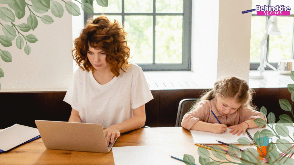 A woman works on a laptop while a young girl beside her draws on paper at a wooden table in a bright room, with green leafy decorations framing the image.