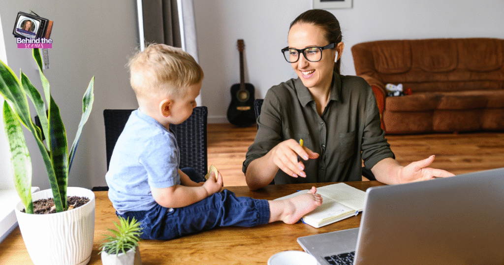A woman works at a laptop, smiling and talking to a young child sitting on the table beside her. The room has plants, a sofa, a guitar, and a notebook and coffee cup on the table.