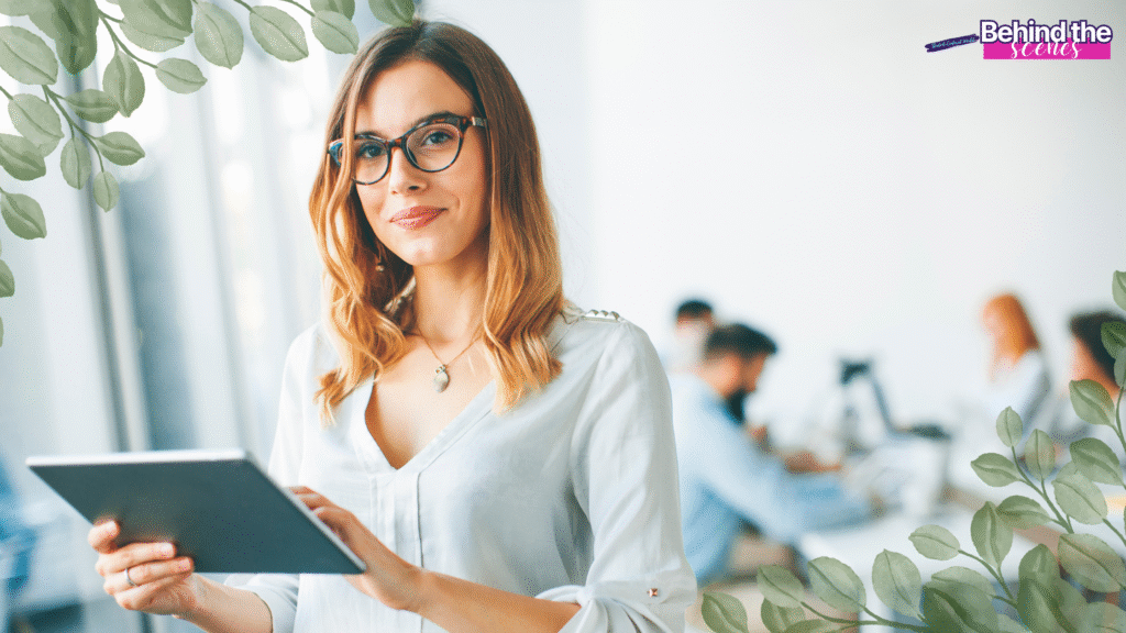 A woman with glasses, holding a tablet and smiling, stands in a bright office—capturing her transition from full time job to entrepreneur. Several people work at desks in the blurred background. Green leaves frame the image and a Behind the Scenes label appears in the top right corner.
