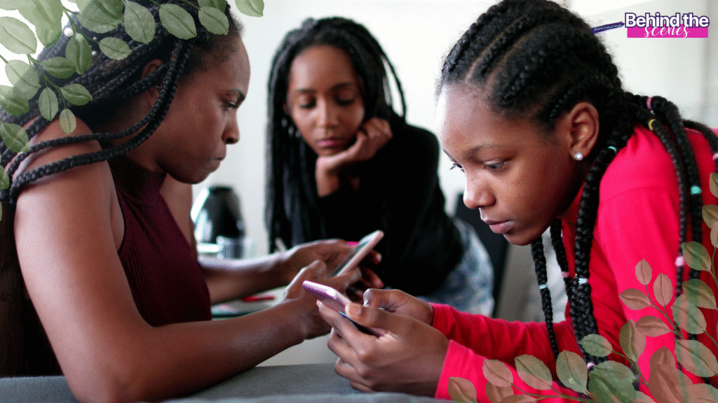 Three young Black women with braided hair sit closely together indoors, focused on their smartphones. Decorative green leaves border the image corners, and a Behind the scenes label appears at the top right.