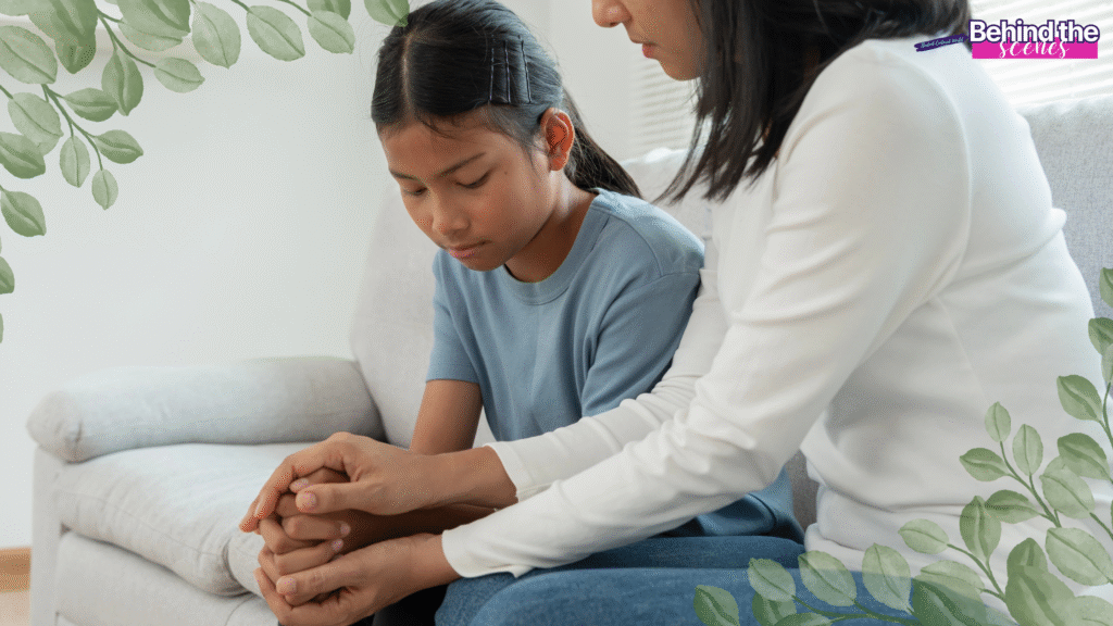A woman sits beside a sad young girl on a couch, holding her hands for comfort. Both appear serious and thoughtful. Green leaves frame the image, and Behind the scenes text is in the top right corner.
