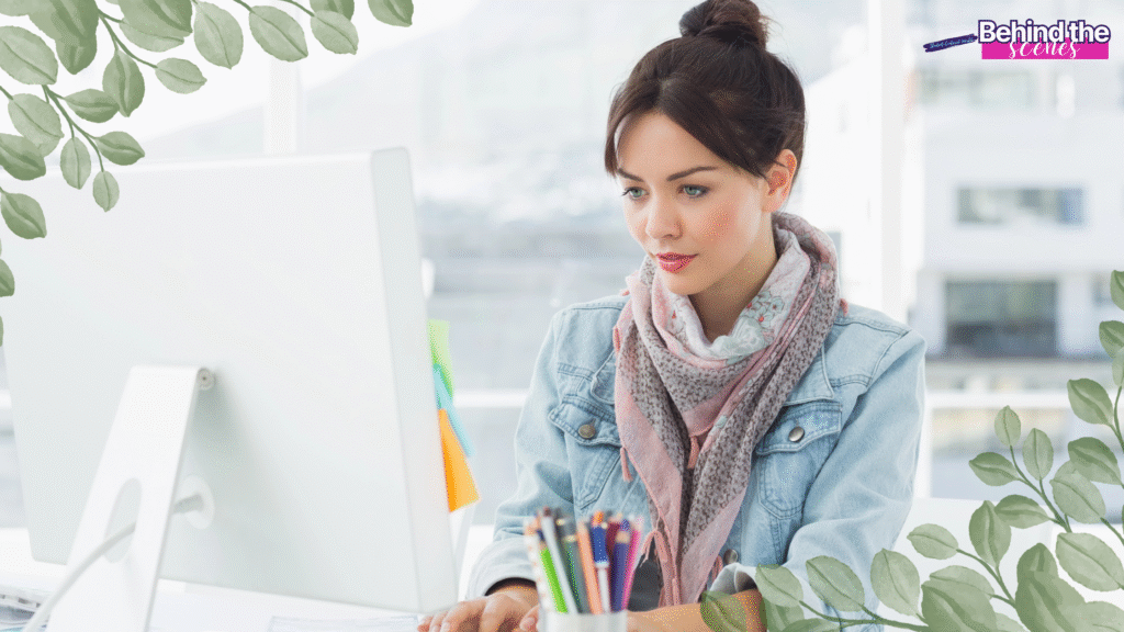 A woman with dark hair in a bun, wearing a scarf and denim jacket, works at a desktop computer in a bright office—highlighting flexible work from home jobs for moms. Green leafy graphics border the image, with a Behind the Scenes label in the top right.