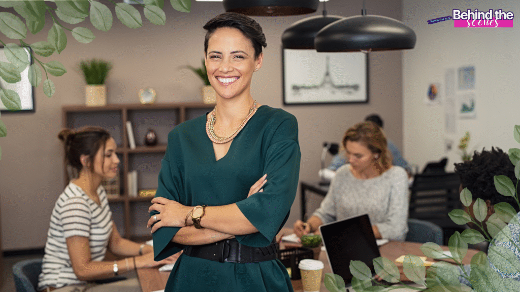 A confident woman in a green dress stands smiling with arms crossed in a modern office, embodying the transition from full time job to entrepreneur. Leaves frame the image, with a “Behind the scenes” label in the top right corner.