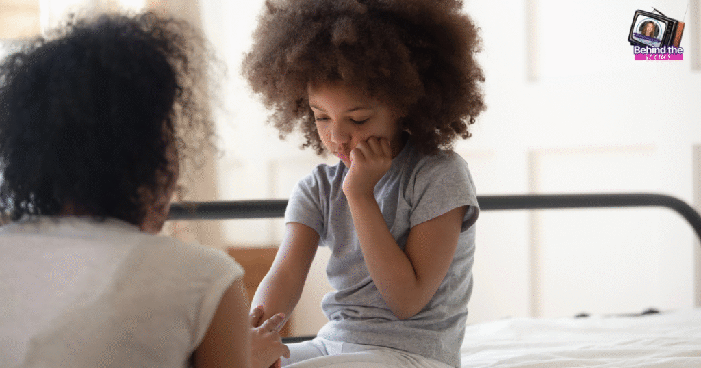 A young girl with curly hair sits on a bed, looking thoughtful and resting her chin on her hand, while an adult, seen from behind, sits nearby, gently holding her other hand.