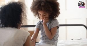 A young girl with curly hair sits on a bed, looking thoughtful and resting her chin on her hand, while an adult, seen from behind, sits nearby, gently holding her other hand.