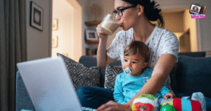 A woman sits on a couch with a baby on her lap, sipping a drink while looking at a laptop—balancing motherhood and the transition from a full-time job to entrepreneur. The cozy room features toys, a pacifier, and framed photos on the walls.