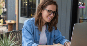 A woman with brown hair and glasses, wearing a denim shirt over a white top, smiles while working on a laptop outdoors—embracing flexible work from home jobs for moms. A small Behind the Scenes logo appears in the top right corner.