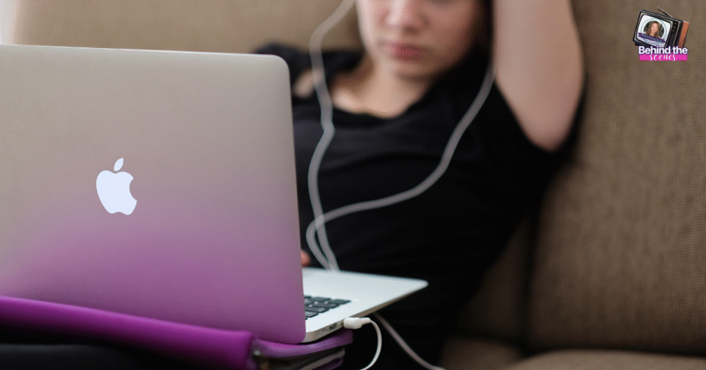 A person in a black shirt sits on a couch with one arm resting above their head, using a silver MacBook laptop and wearing white earphones. A small Behind the Reality logo appears in the top right corner.