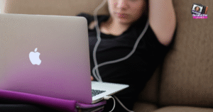 A person in a black shirt sits on a couch with one arm resting above their head, using a silver MacBook laptop and wearing white earphones. A small Behind the Reality logo appears in the top right corner.