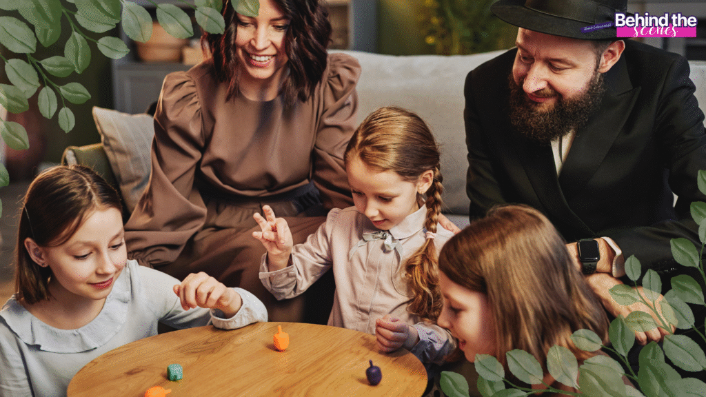 A family with three children sits around a wooden table, smiling and playing with colorful dreidels. Leaves frame the image, and the words Behind the scenes appear at the top right corner. holiday overwhelm kids