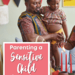 A man holds a young girl near a table with lit red candles. Both wear patterned clothes. A sign in front reads, Parenting a Sensitive Child throughout the holidays. Colorful banners hang on the wall in the background.