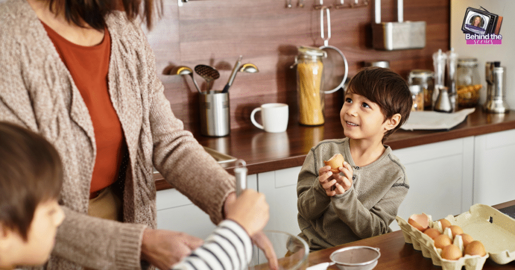 A young boy smiles and holds an egg while standing at a kitchen counter with an adult and another child, preparing to cook together. Kitchen utensils and food jars are visible in the background.
