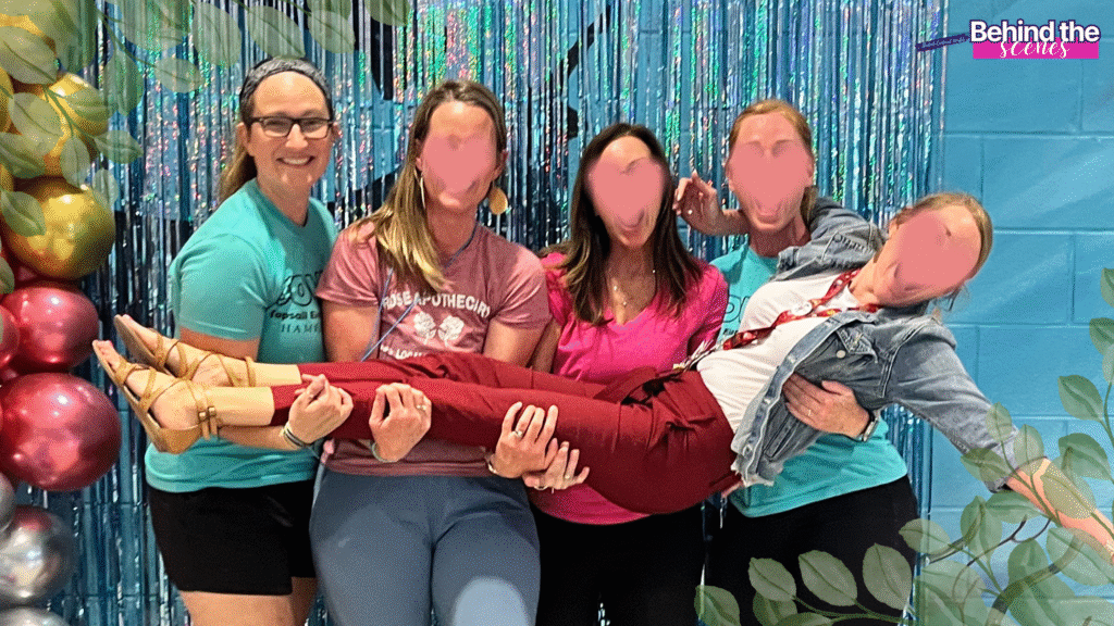 Five women pose in front of a festive, sparkly backdrop with balloons. Four are standing and smiling while holding up the fifth woman horizontally across them. A “Behind the Scenes” sign is in the top right corner.