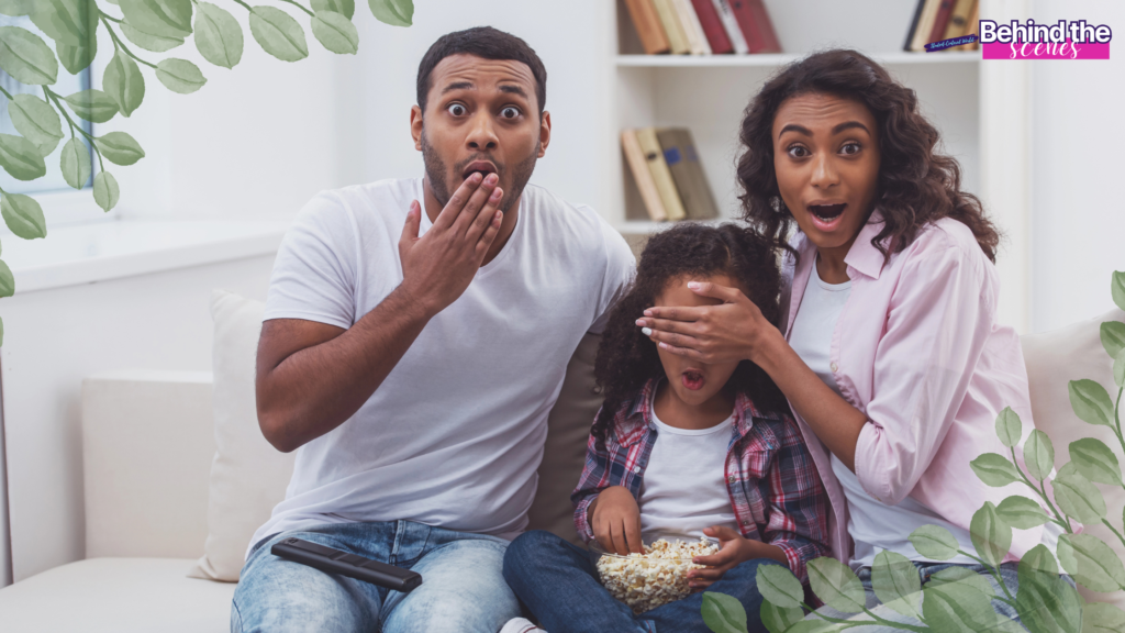 A surprised man and woman sit on a couch with a young girl between them; the woman covers the girl’s eyes while she holds popcorn. All three have shocked expressions. Green leaves frame the image, and there’s a Behind the scenes label.