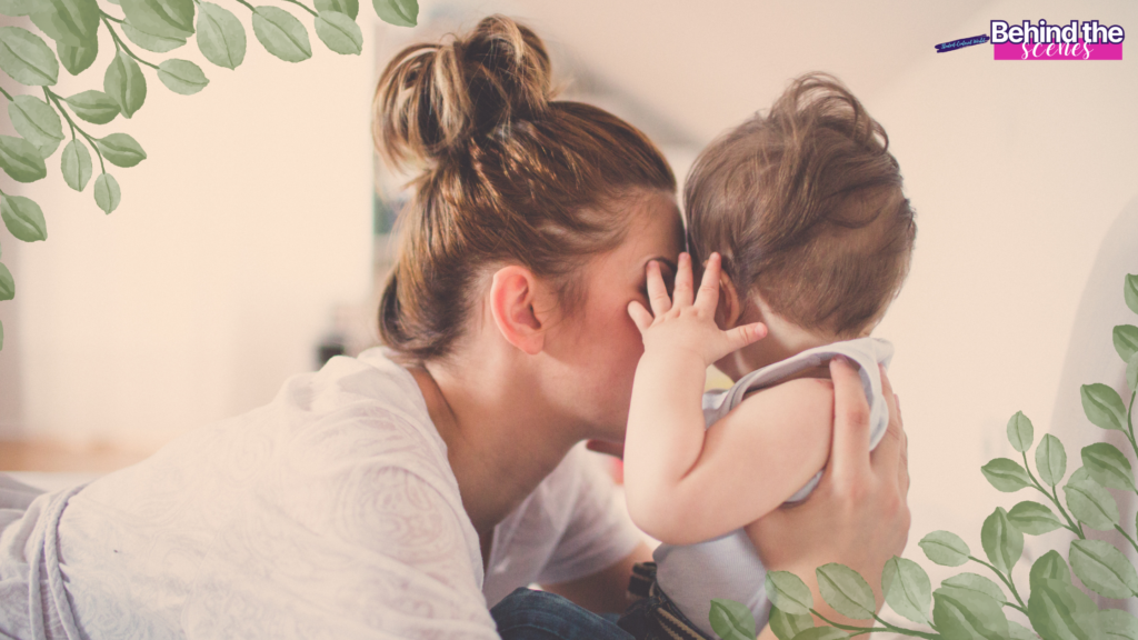 A woman gently hugs and whispers to a baby indoors, surrounded by soft, leafy decorations. The scene looks warm and intimate, with Behind the Scenes written in the top right corner. Finding purpose after motherhood.
