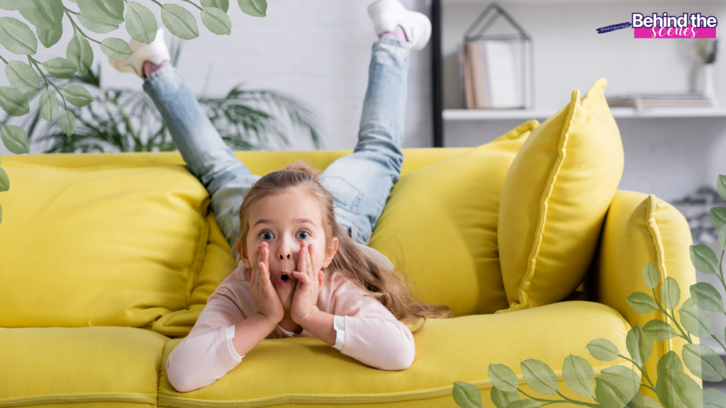 A young child lies on a bright yellow couch with legs up in the air, holding their face in surprise. Green leafy plants frame the image, capturing a moment that shows how to make home feel safe. A Behind the scenes label appears in the top right corner.