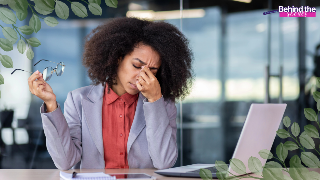 A woman in a blazer sits at a desk with a laptop, holding her glasses in one hand and pinching the bridge of her nose, visibly stressed—a moment of career burnout recovery. Green leaves frame the image, and Behind the scenes is written in the corner.