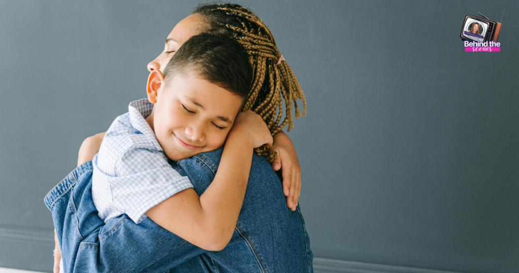 A young boy with short hair smiles with his eyes closed as he hugs an adult in a denim jacket, showing how moments like these make home feel safe. The background is a plain dark wall with a small “Behind the Scenes” sticker in the corner.