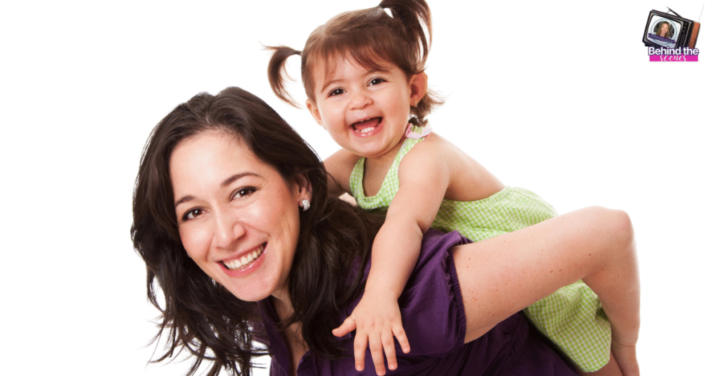 A smiling woman gives a piggyback ride to a happy young girl in a green dress; both are laughing against a white background. A small TV graphic with text Behind the Media appears in the top right corner. Reinventing yourself after kids