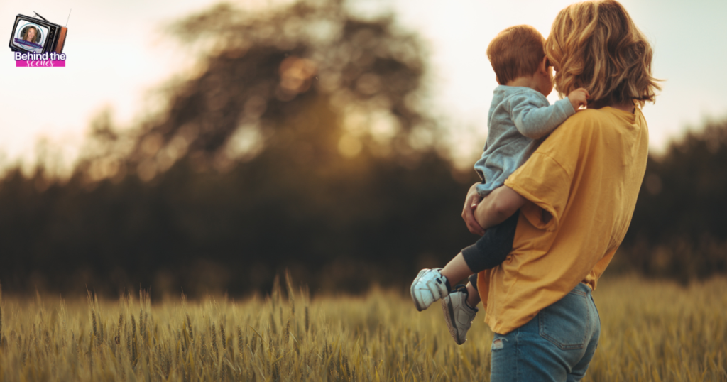 A woman in a yellow shirt stands in a field, holding a young child as they face away from the camera toward blurred trees at sunset. A small Behind the Reality TV logo is in the top left corner.