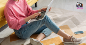 A person in a pink sweatshirt and jeans sits on the floor using a laptop, perhaps researching career burnout recovery. A smartphone is nearby, with a yellow couch, patterned rug, and a Behind the Scenes sticker in the top right corner.