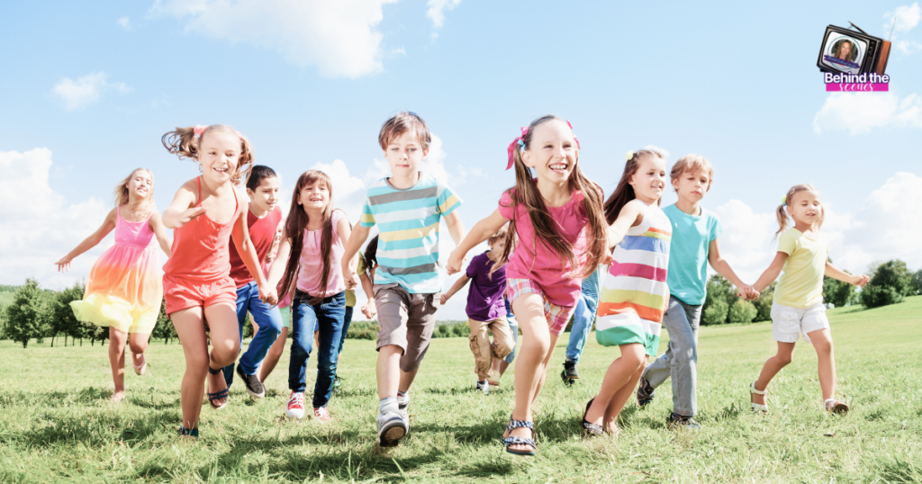 A group of happy children in colorful clothes run together on a grassy field under a bright blue sky with scattered clouds. In the top right corner, there is a logo that says Behind the Victory.